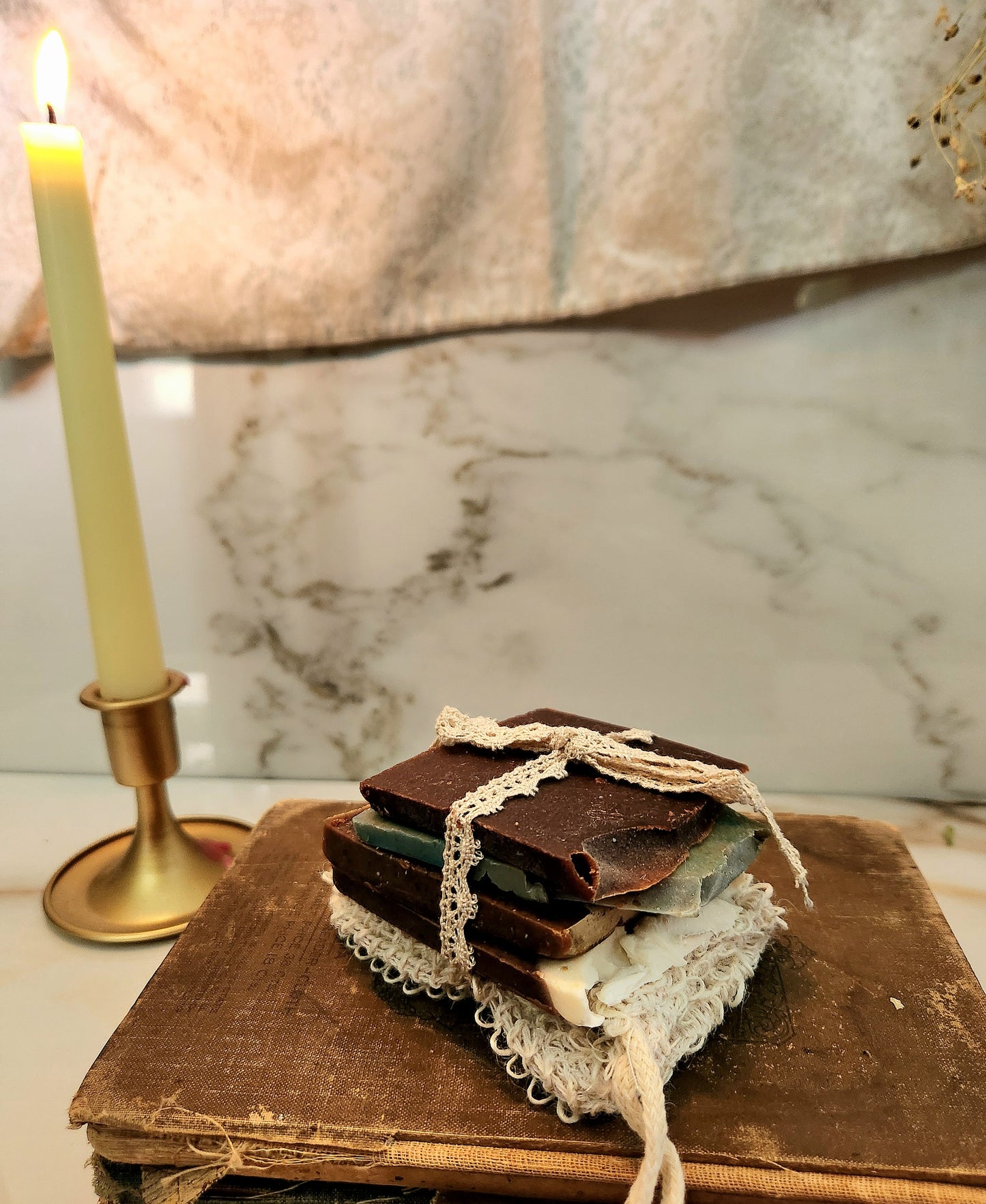 A stack of soap bars tied with cream lace ribbon, resting on an old book next to a lit candle in a brass holder, set against a marble-patterned background.