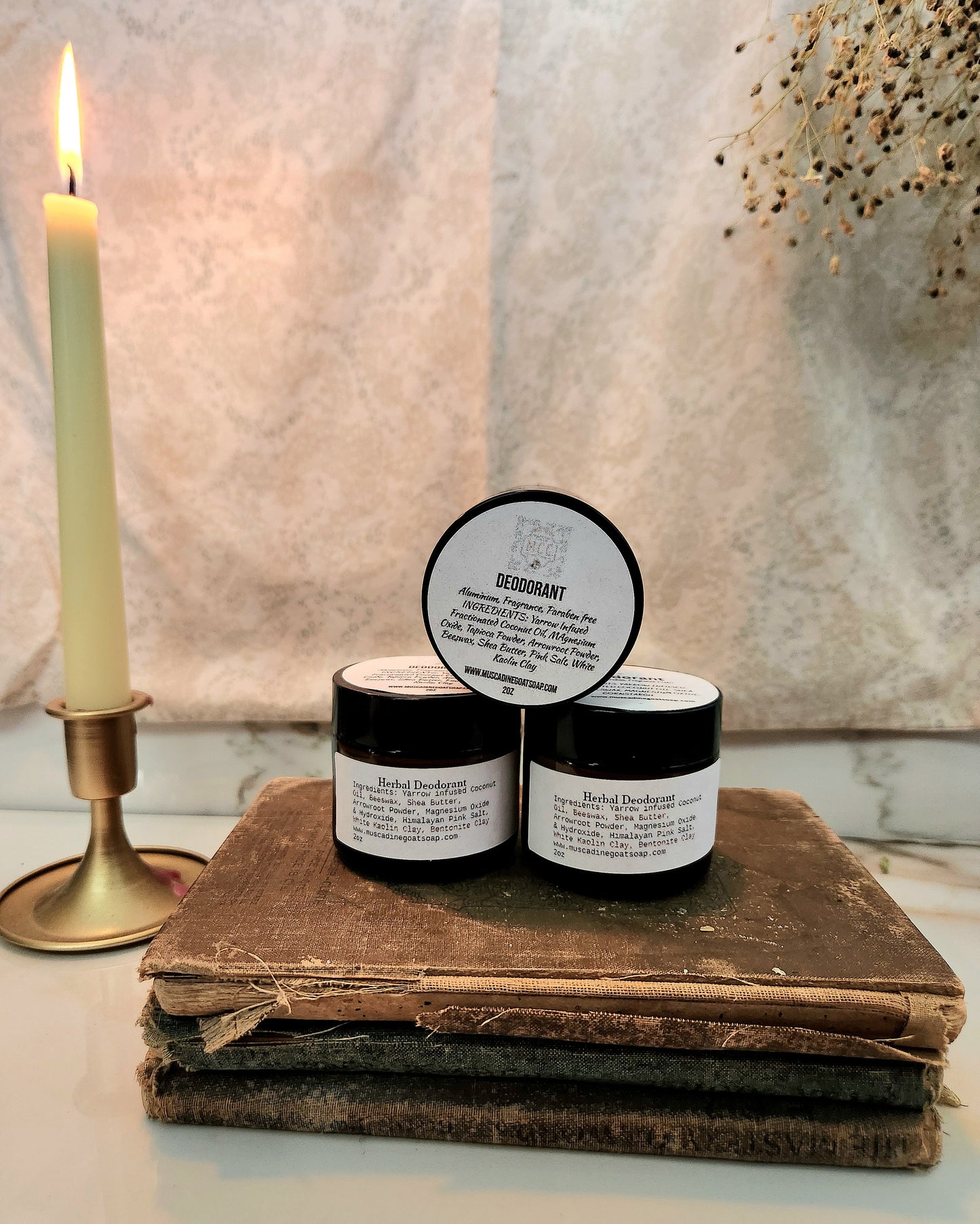 Two small jars of Herbal deodorant, displayed on a stack of old books next to a lit candle.