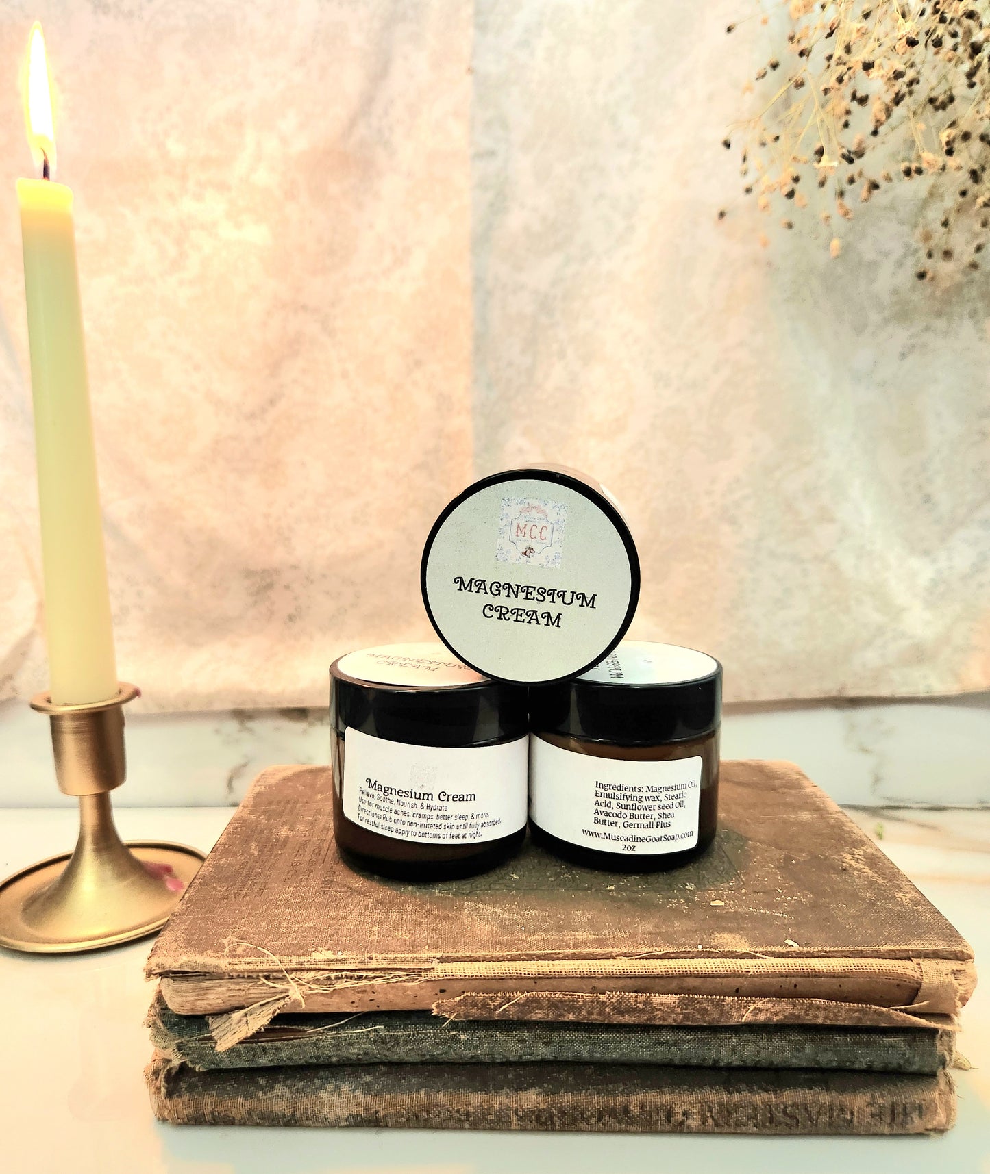 Two small glass amber jars labeled 'MAGNESIUM CREAM' with a black lid on top, displayed on a stack of old books next to a lit candle against a textured beige backdrop.