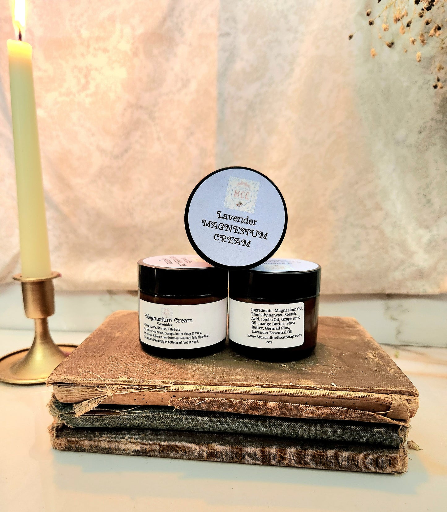 Two small black jars of cream labeled Lavender Magnesium Cream, styled on a stack of old books with a lit candle in the background.