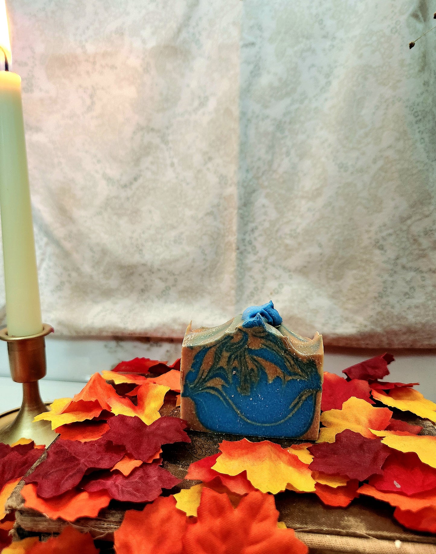 A rectangular bar of handmade soap with a blue and orange pattern sits on a wooden surface surrounded by red, orange, and yellow autumn leaves. A lit white candle in a brass holder is visible on the left, with a textured light fabric backdrop behind.