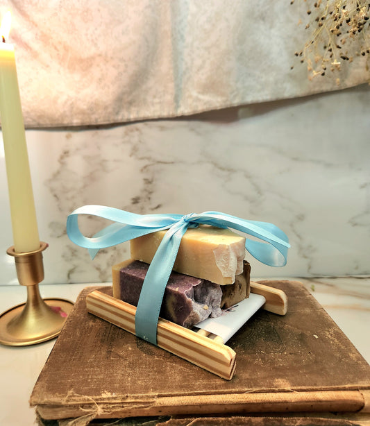 A set of artisanal soap bars wrapped in light blue ribbon and presented on a small wooden soap deck, styled on top of an old book next to a lit candle.