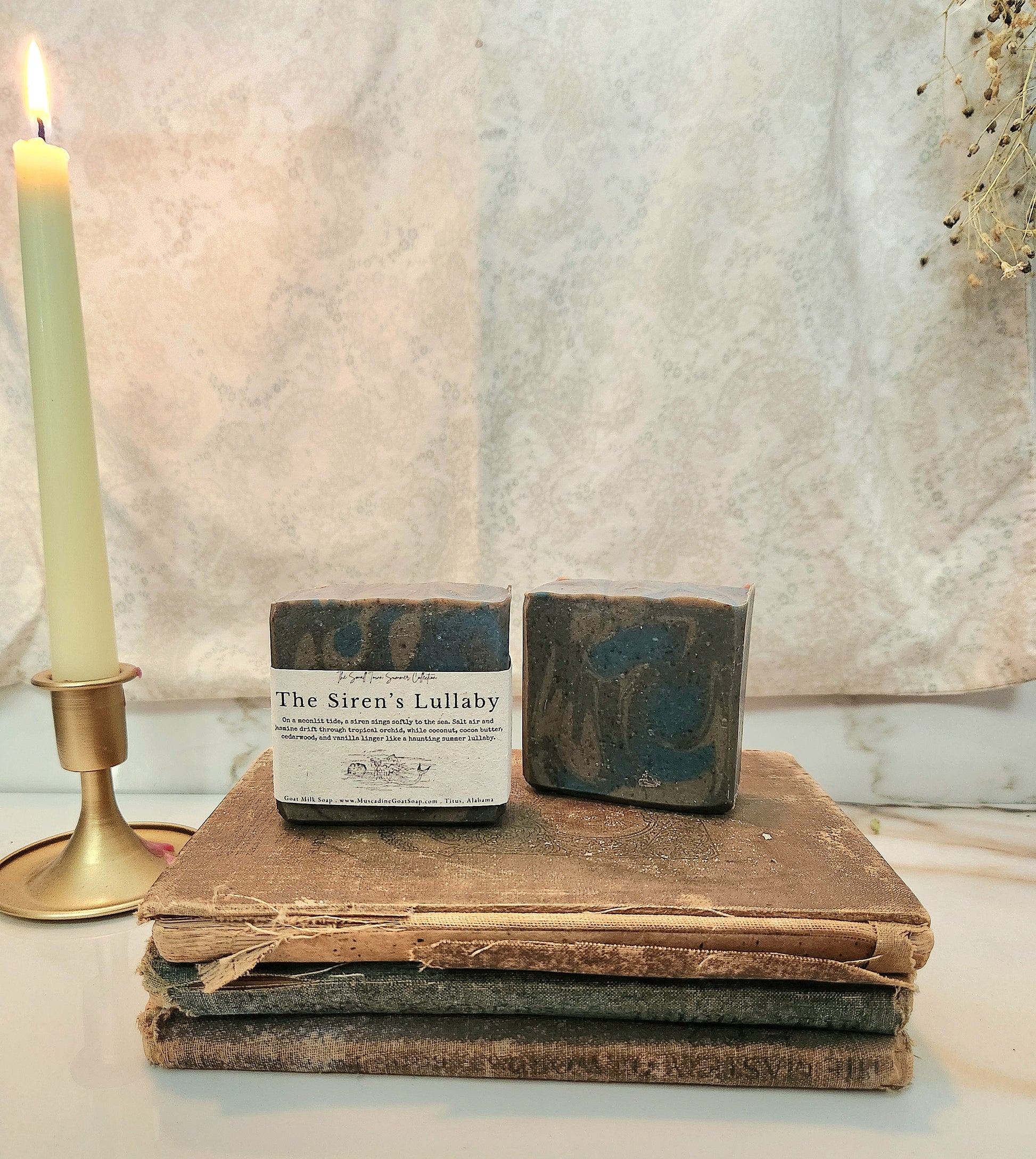 Two artisanal soap bars labeled 'The Siren's Lullaby' displayed on a stack of vintage books, with a lit candle in a brass holder on the left and a textured beige backdrop behind.