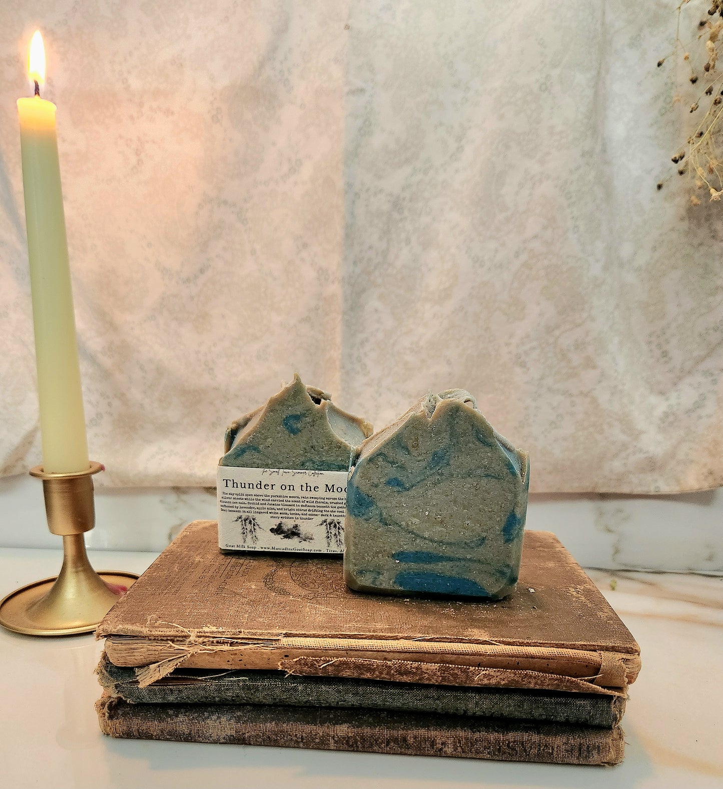 Two handmade artisan soap bars with a blue and gray marbled pattern rest on a stack of vintage brown books next to a lit cream-colored candle in a brass holder.