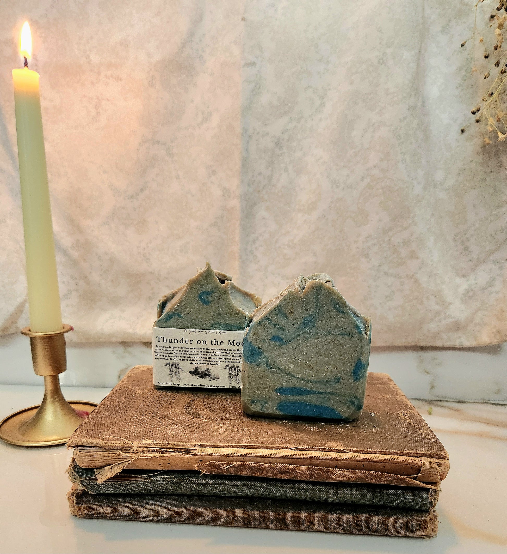 Two handmade artisan soap bars with a blue and gray marbled pattern rest on a stack of vintage brown books next to a lit cream-colored candle in a brass holder.