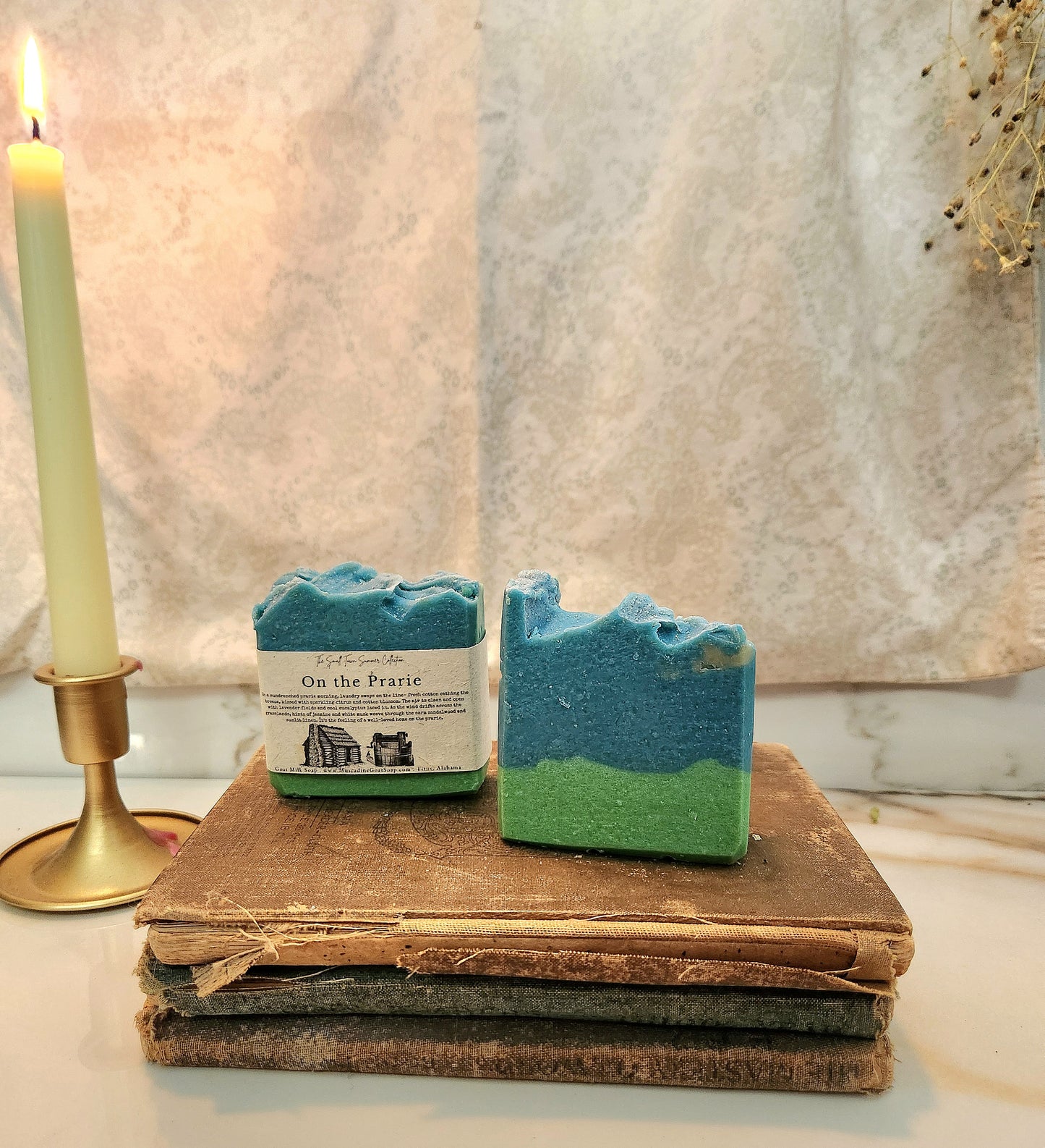 Two handmade soap bars labeled 'On the Prairie' are displayed on a stack of vintage books next to a lit candle in a brass holder. The soap features a blue and green swirled pattern, set against a rustic background with a textured beige fabric backdrop.