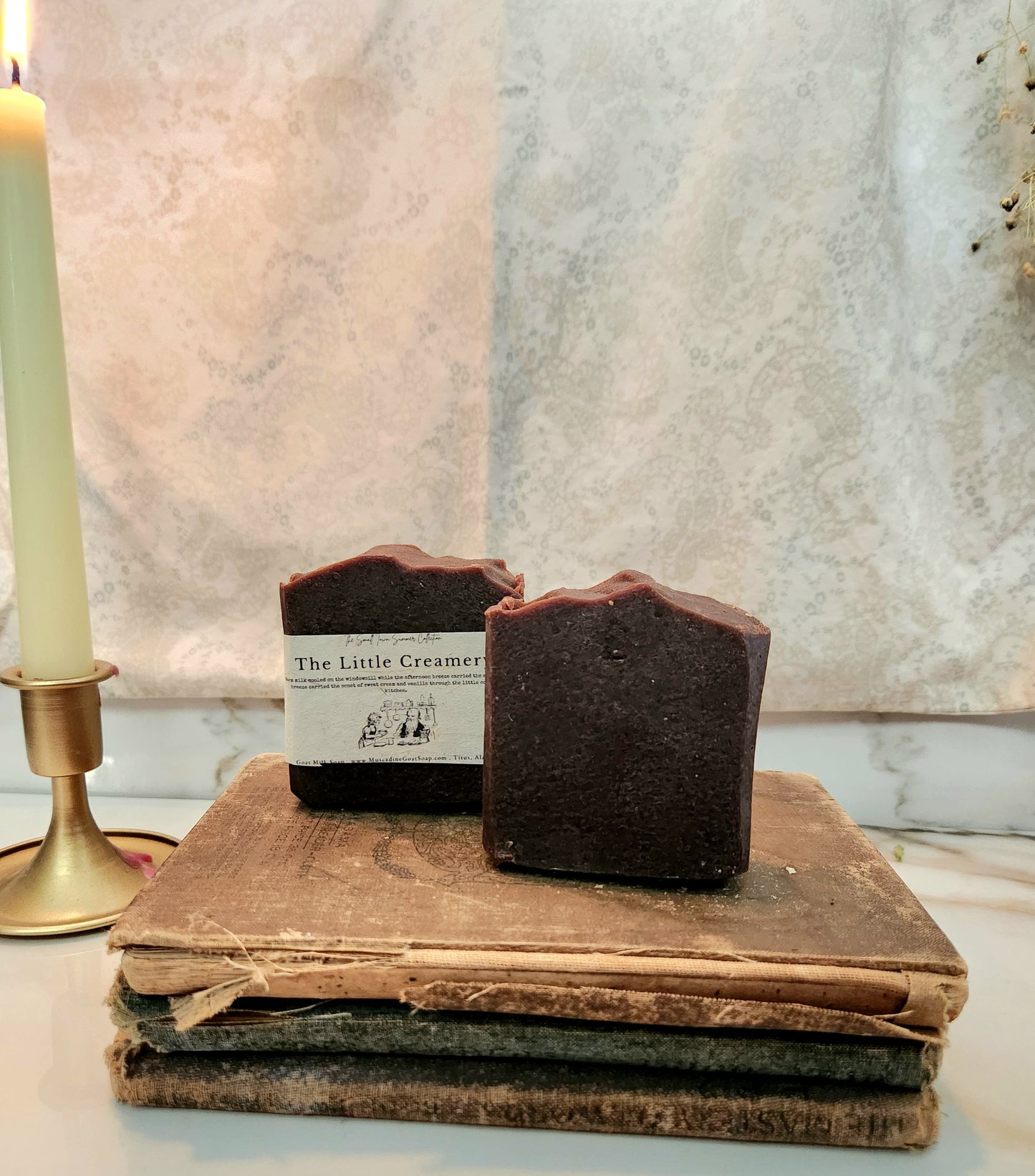Two dark chocolate-colored handmade soap bars labeled 'The Little Creamery' resting on a stack of vintage books, with a lit candle in a brass holder on the left.