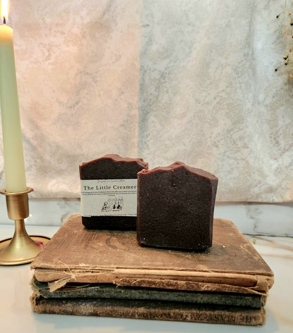 Two dark chocolate-colored handmade soap bars labeled 'The Little Creamery' resting on a stack of vintage books, with a lit candle in a brass holder on the left.