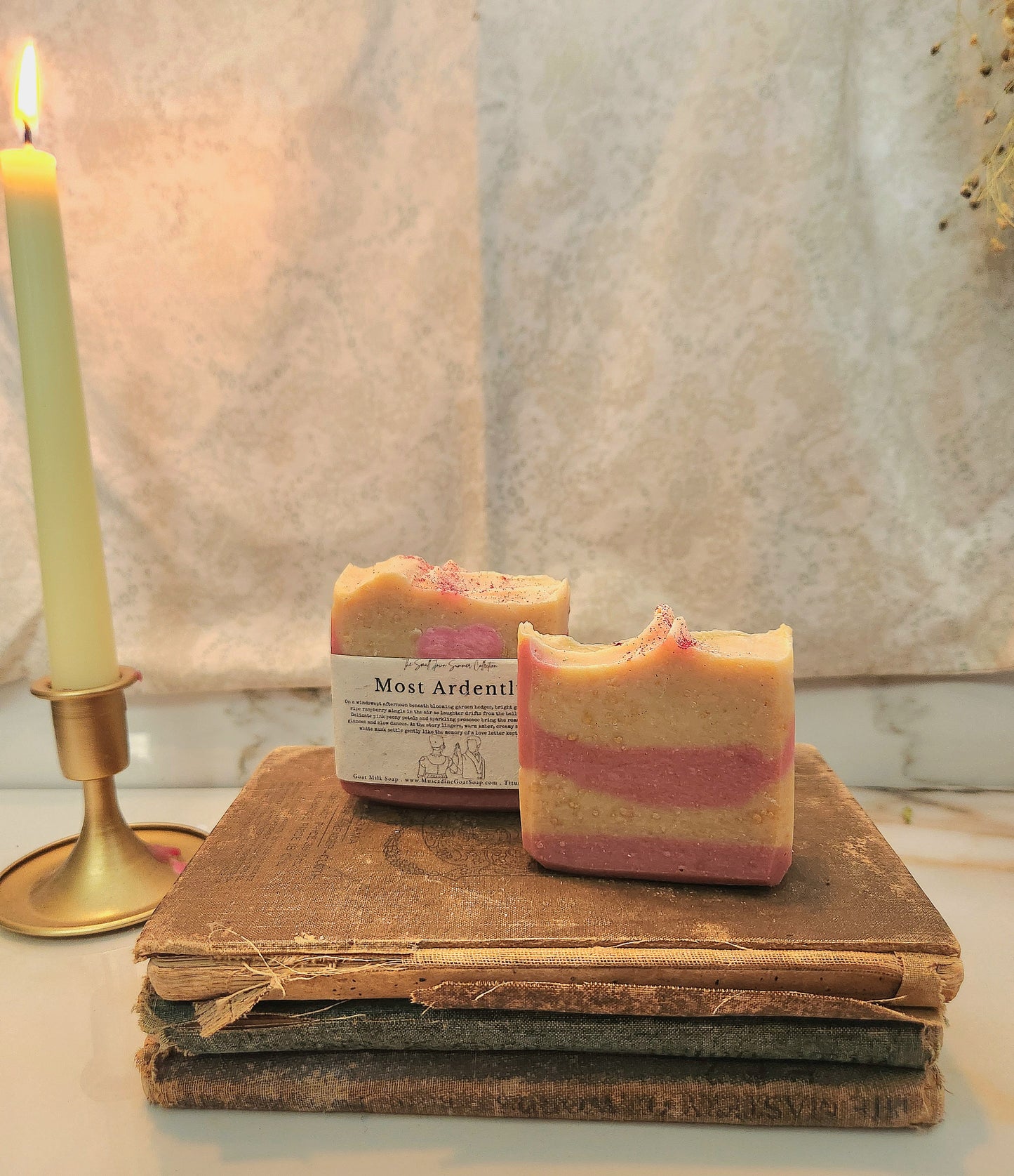 Two rectangular handmade soap bars with a pink and cream marbled pattern rest on a stack of vintage books next to a lit candle in a brass holder. The soap bar in the foreground has a seed-embedded paper label reading 'Most Ardently'.