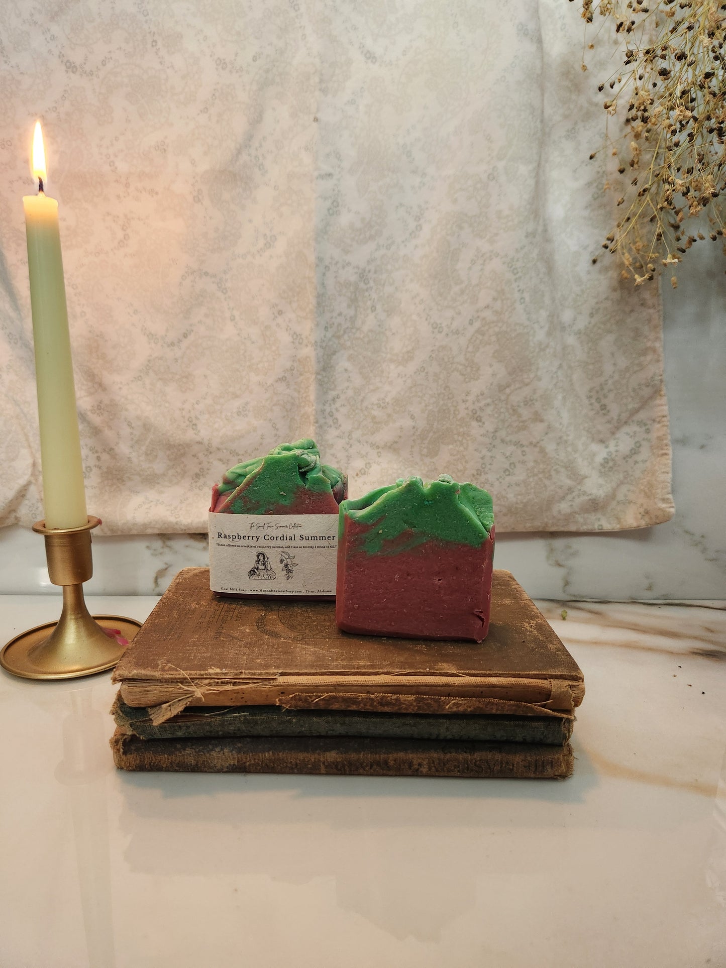 A handmade soap bar labeled Raspberry Cordial Summer, displayed on a stack of old books next to a lit candle in a brass holder, set against a neutral draped fabric background.