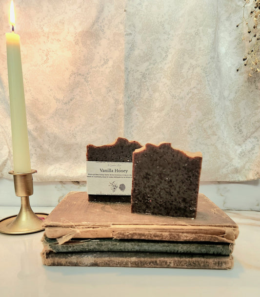Two rustic handmade soap bars labeled 'Vanilla History' displayed on a stack of vintage books, with a lit candle in a brass holder to the left against a textured light backdrop.