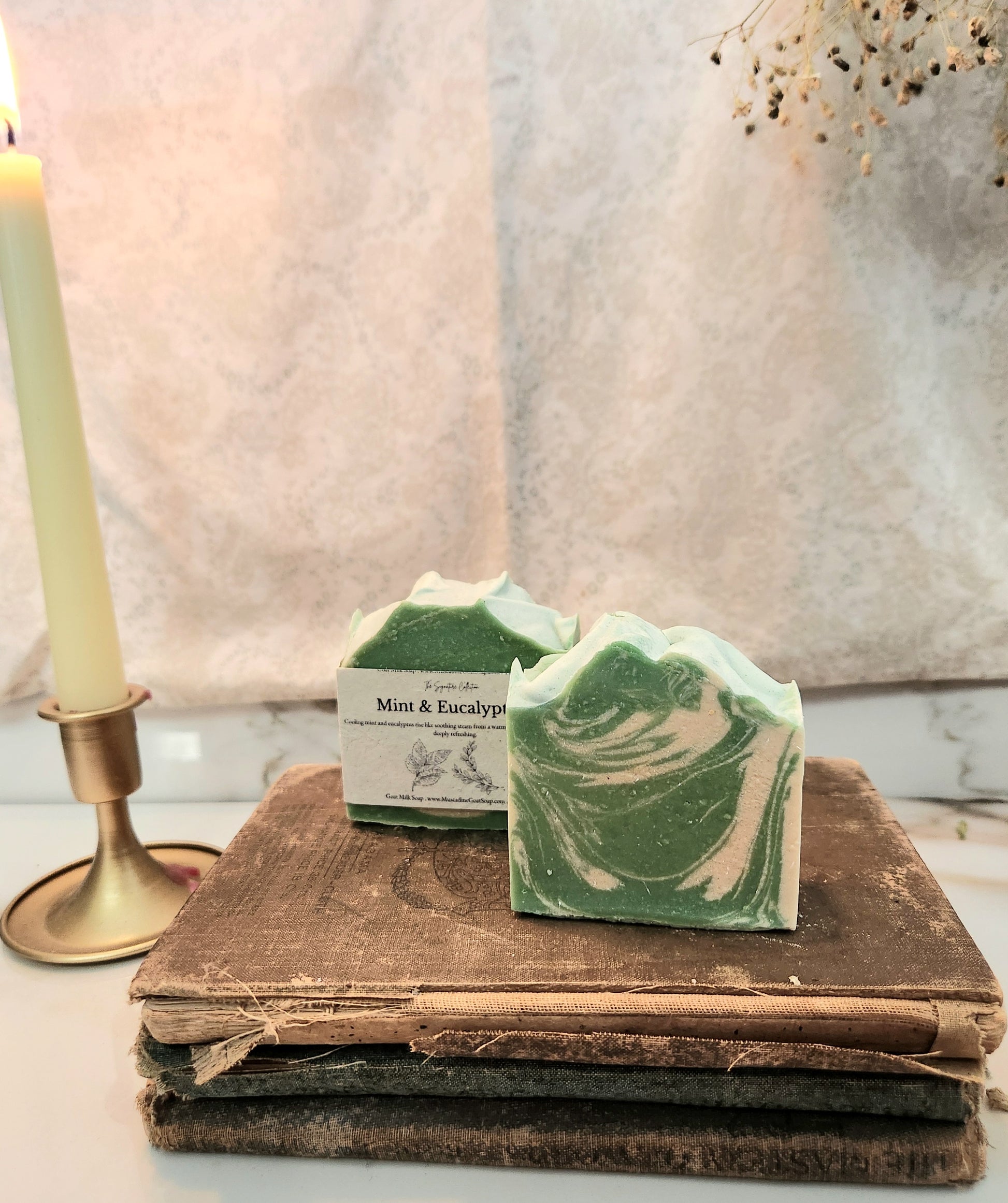Two handmade soap bars with green and white marbled swirls are displayed on a stack of vintage wooden books next to a lit cream-colored candle in a brass holder. The background features a soft, draped beige fabric.