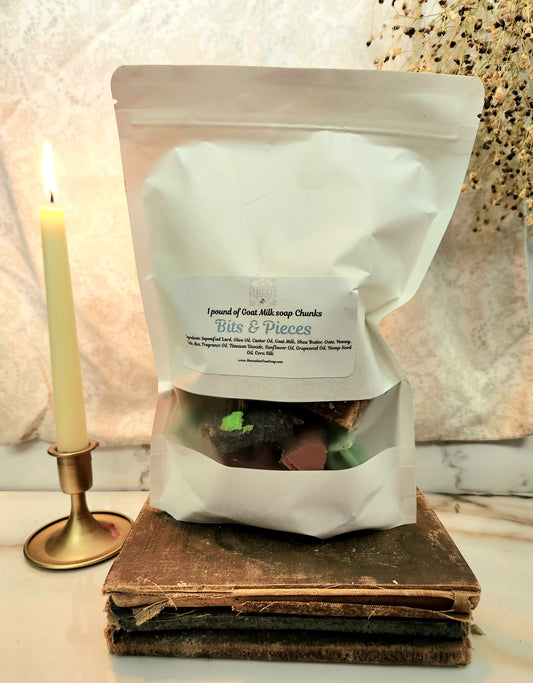 A white stand-up pouch labeled '1 pound of goat milk soap 'Bits & Pieces' sits on stacked vintage books next to a lit candle in a brass holder, with a textured beige background.