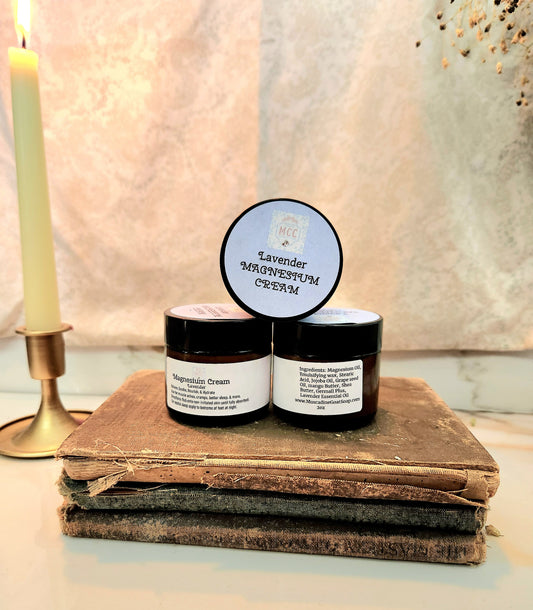 Two small black jars of cream labeled Lavender Magnesium Cream, styled on a stack of old books with a lit candle in the background.