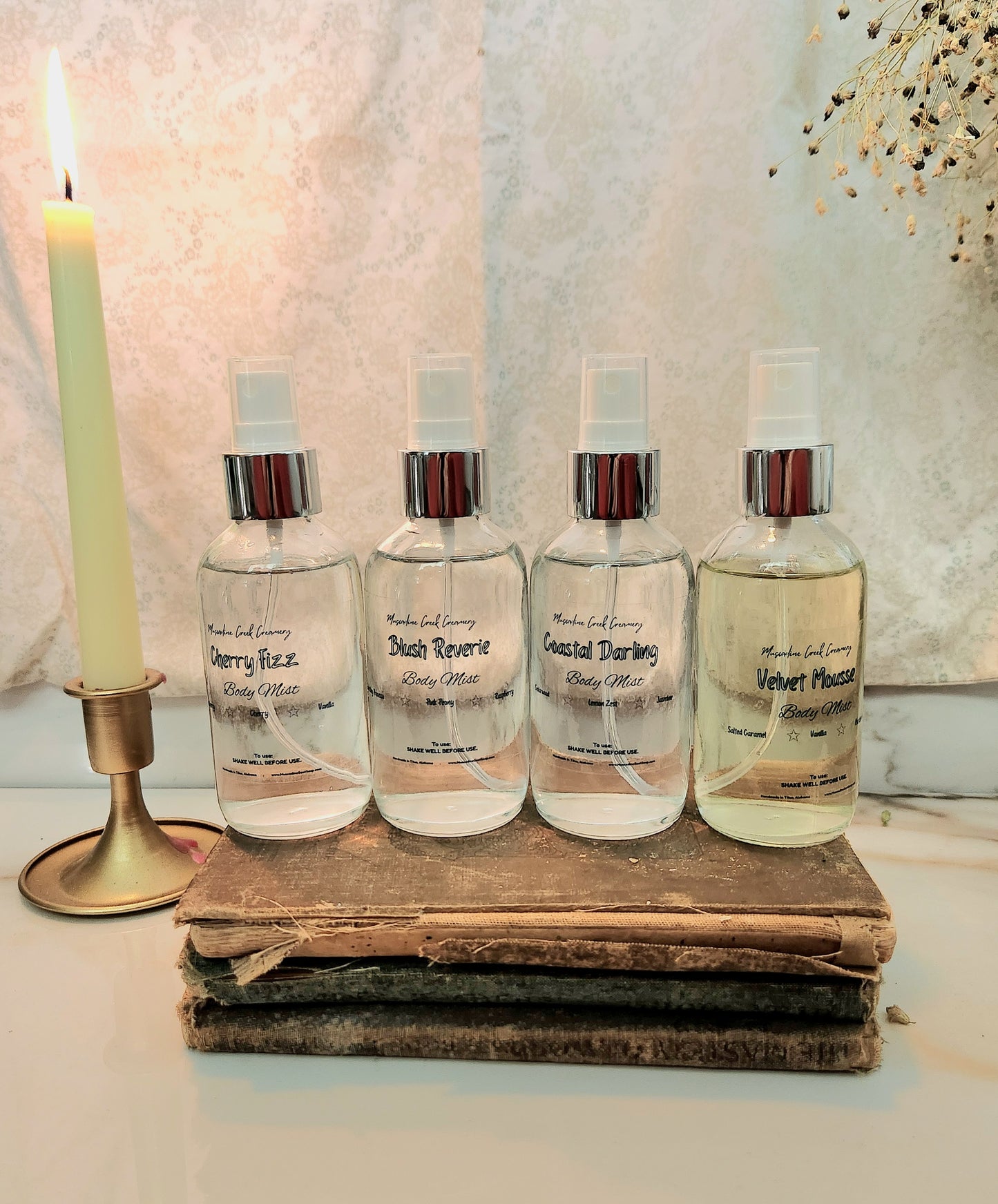 Four clear spray bottles with silver caps arranged on a stack of old books next to a lit candle, against a textured light background.
