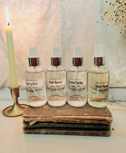 Four clear spray bottles with silver caps arranged on a stack of old books next to a lit candle, against a textured light background.