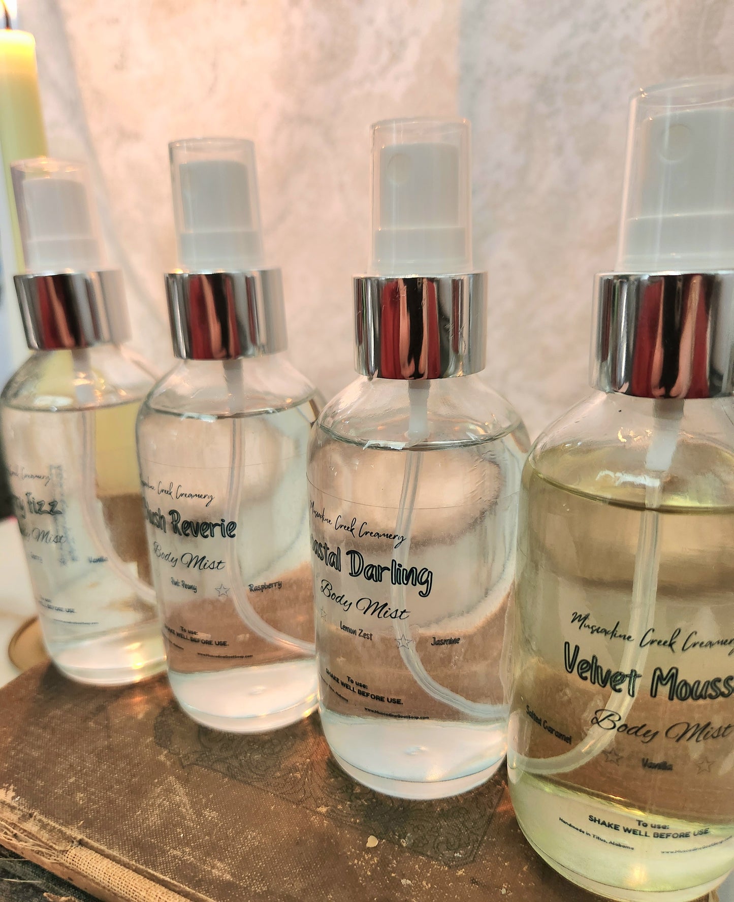 Four clear glass spray bottles of Body mist with silver collars and white sprayers, lined up on a rustic book pile. The labels show scents including Cherry Fizz, Velvet Mousse, Coastal Darling, and Blush Reverie, & Velvet Mousse.
