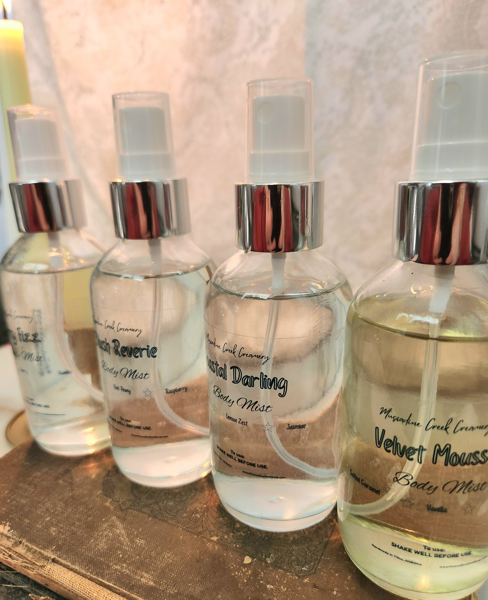 Four clear glass spray bottles of Body mist with silver collars and white sprayers, lined up on a rustic book pile. The labels show scents including Cherry Fizz, Velvet Mousse, Coastal Darling, and Blush Reverie, & Velvet Mousse.