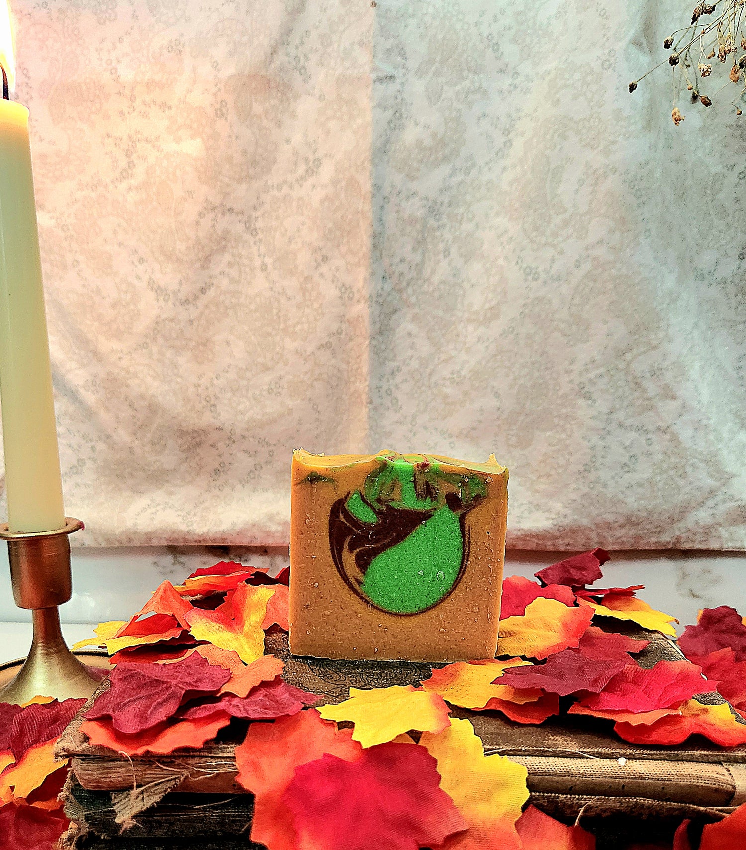 A rectangular bar of handmade soap with a yellow base and a green and dark red swirl pattern, displayed on a rustic wooden surface surrounded by red, orange, and yellow autumn leaves. A lit candle in a brass holder is visible on the left, with a textured light gray fabric backdrop behind.