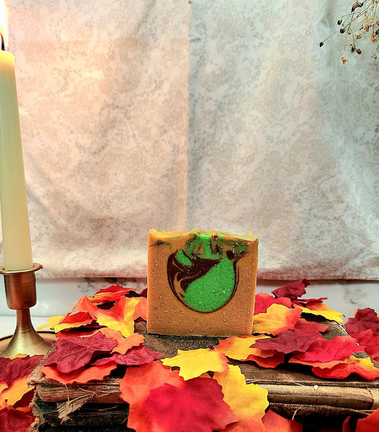 A rectangular bar of handmade soap with a yellow base and a green and dark red swirl pattern, displayed on a rustic wooden surface surrounded by red, orange, and yellow autumn leaves. A lit candle in a brass holder is visible on the left, with a textured light gray fabric backdrop behind.
