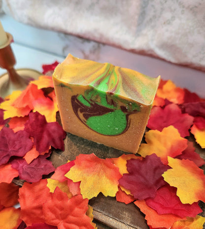 A rectangular bar of handmade soap with a marbled pattern of green, yellow, and dark red sits on a bed of colorful artificial autumn leaves.