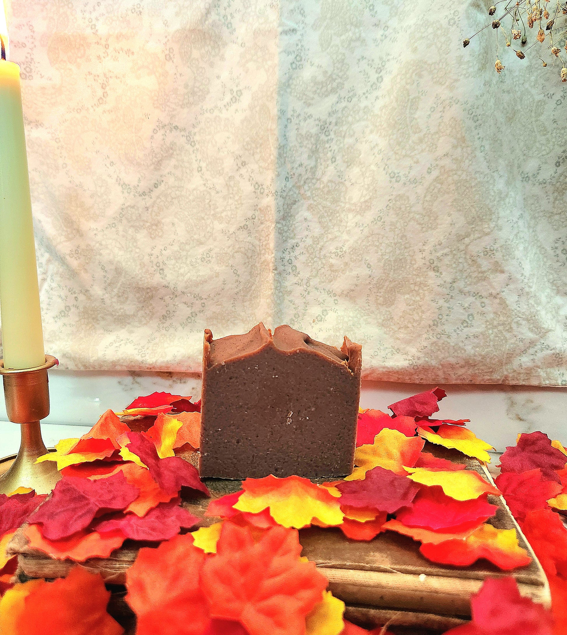 A rectangular bar of dark brown soap bar standing upright on a wooden board surrounded by red, orange, and yellow autumn leaves, with a candle in a brass holder on the left and a patterned fabric backdrop behind.
