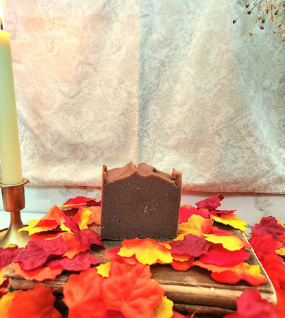 A rectangular bar of dark brown soap bar standing upright on a wooden board surrounded by red, orange, and yellow autumn leaves, with a candle in a brass holder on the left and a patterned fabric backdrop behind.