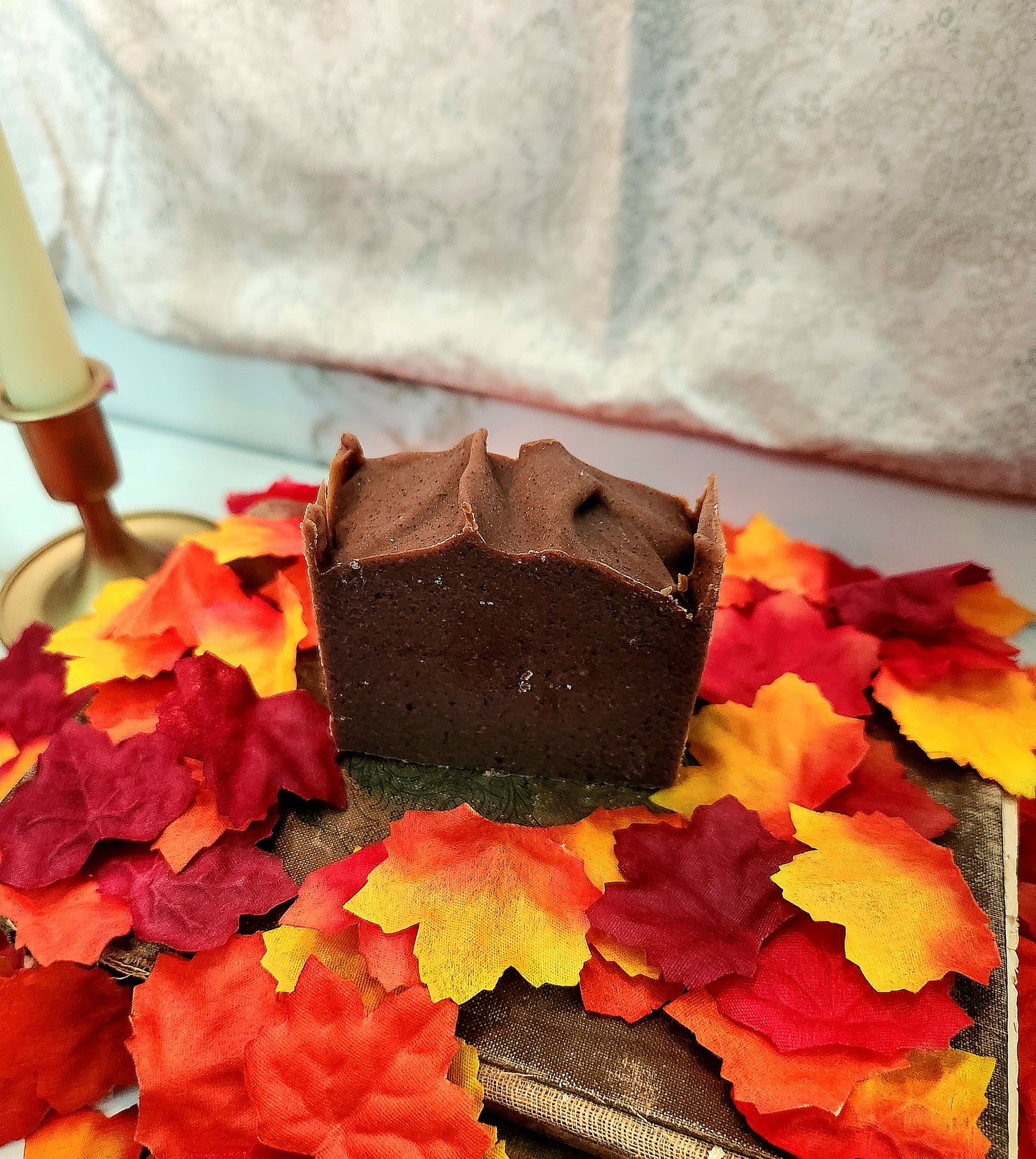 A square bar of dark chocolate handcrafted soap sits on a tray surrounded by colorful autumn leaves, with a candle and soft fabric in the background.