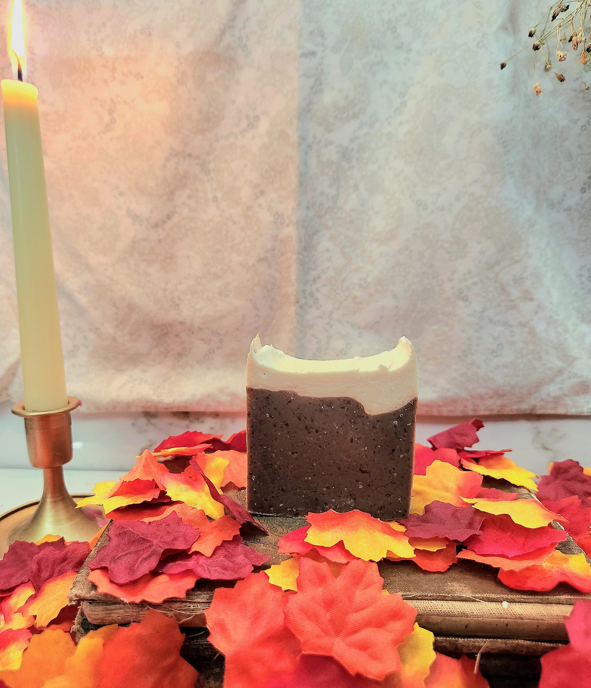 A rectangular handmade soap bar with a dark brown speckled body and a white frosted top, styled on a wooden surface surrounded by red, orange, and yellow autumn leaves, with a lit candle in the background.
