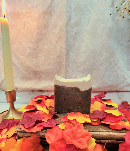 A rectangular handmade soap bar with a dark brown speckled body and a white frosted top, styled on a wooden surface surrounded by red, orange, and yellow autumn leaves, with a lit candle in the background.
