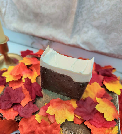 A rectangular bar of soap with a dark brown speckled body and a thick white top layer, styled on a tray surrounded by colorful autumn leaves against a soft neutral background.