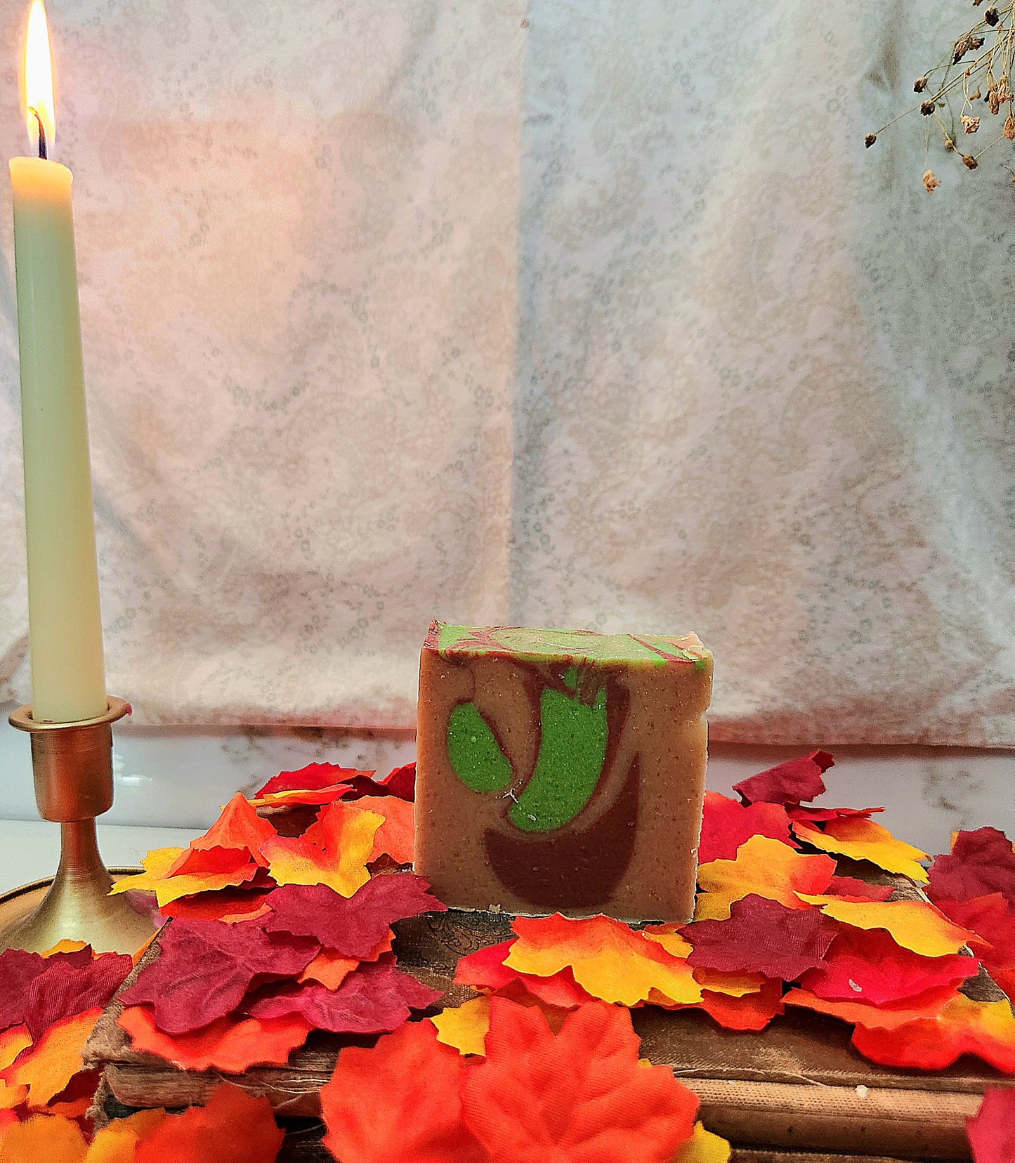 A rectangular bar of handmade soap with a brown base and green and red leaf-like swirls, displayed on a wooden surface scattered with red, orange, and yellow artificial leaves. A lit white candle in a brass holder stands to the left, with a soft beige fabric backdrop behind.