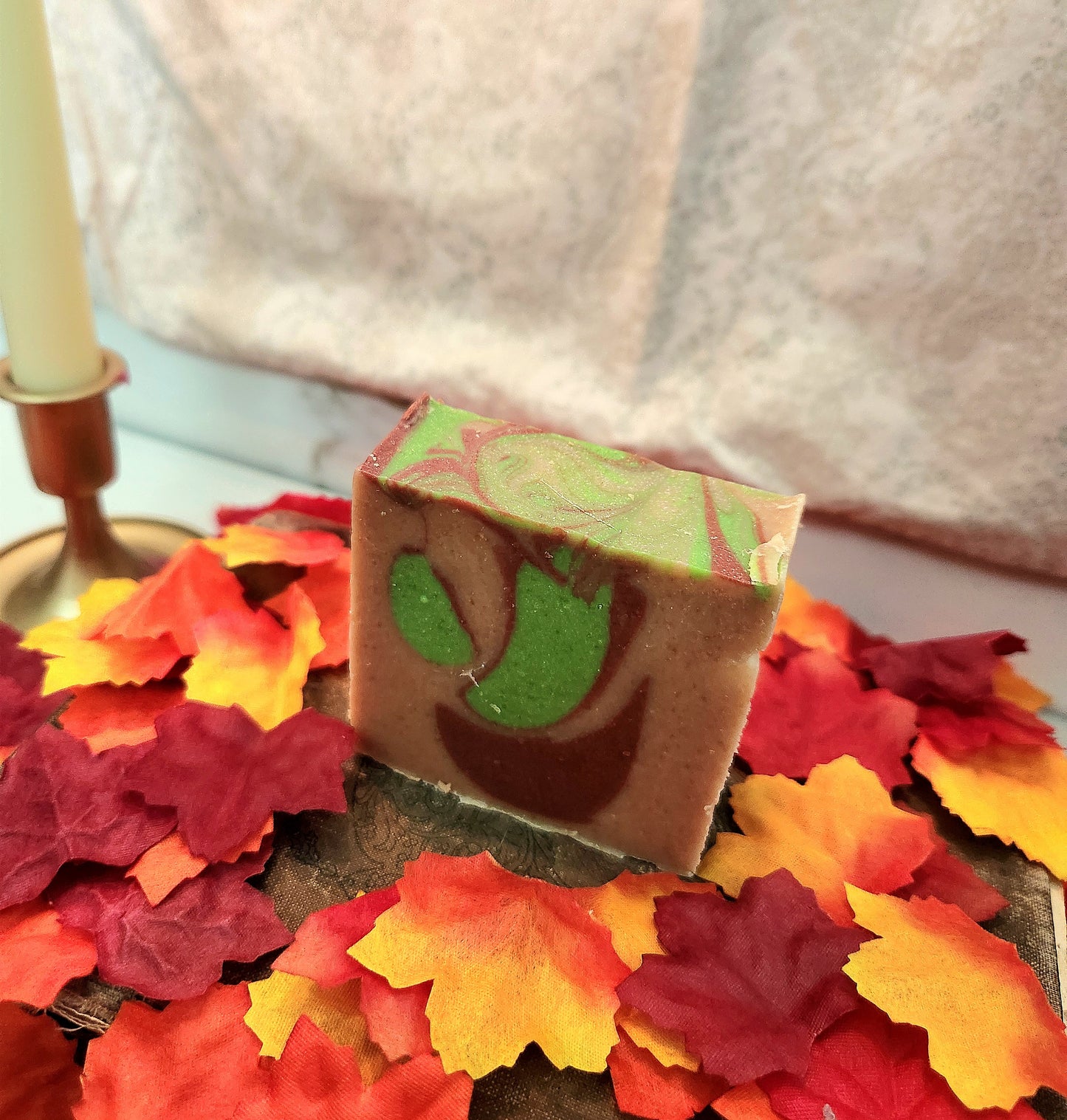 A rectangular bar of artisanal soap with a brown base, green and red marbled top, and a green leaf motif on the front, displayed on a tray surrounded by colorful autumn leaves.