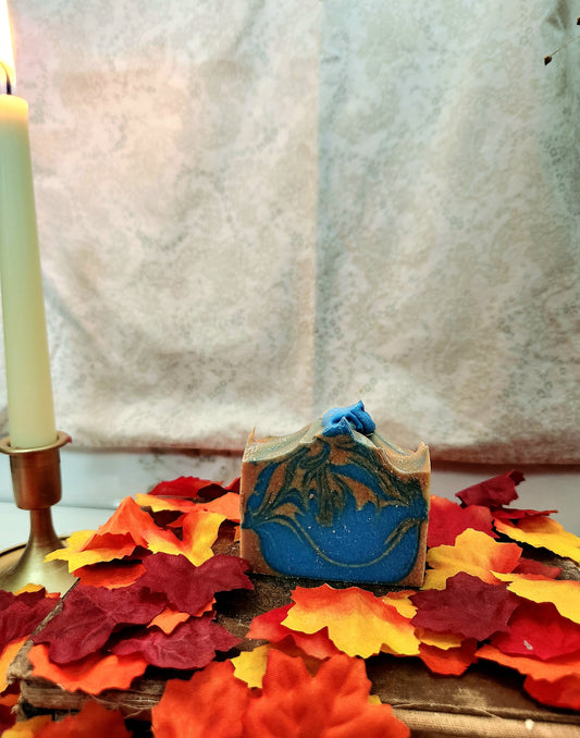 A rectangular bar of handmade soap with a blue and orange pattern sits on a wooden surface surrounded by red, orange, and yellow autumn leaves. A lit white candle in a brass holder is visible on the left, with a textured light fabric backdrop behind.