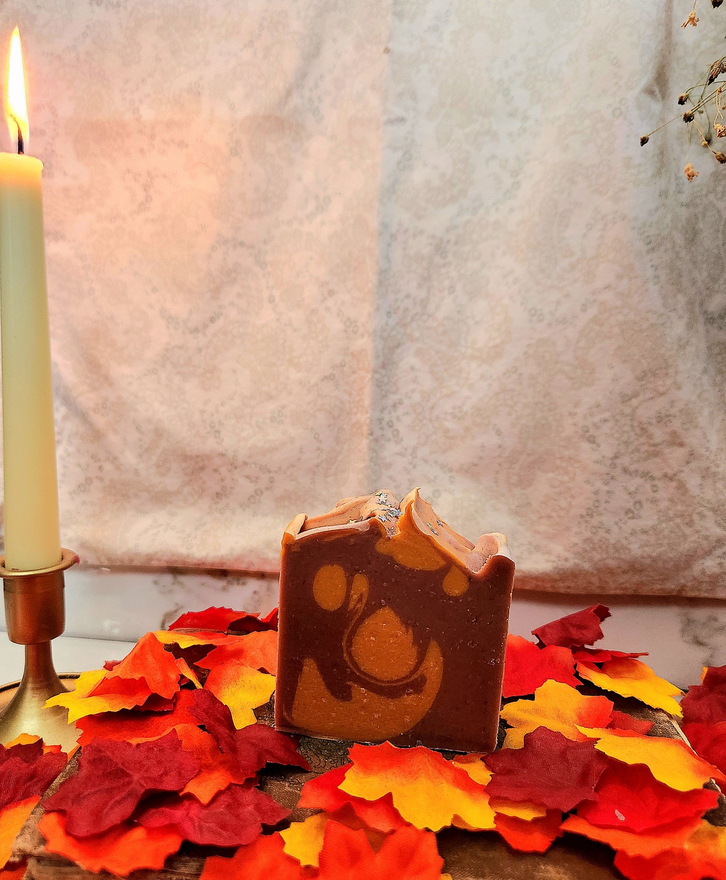 A handmade rectangular bar of soap with a swirled red and orange pattern sits on a wooden surface scattered with red, orange, and yellow autumn leaves. A lit candle in a brass holder is visible on the left, with a soft neutral fabric backdrop behind.