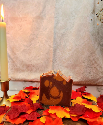 A handmade rectangular bar of soap with a swirled red and orange pattern sits on a wooden surface scattered with red, orange, and yellow autumn leaves. A lit candle in a brass holder is visible on the left, with a soft neutral fabric backdrop behind.