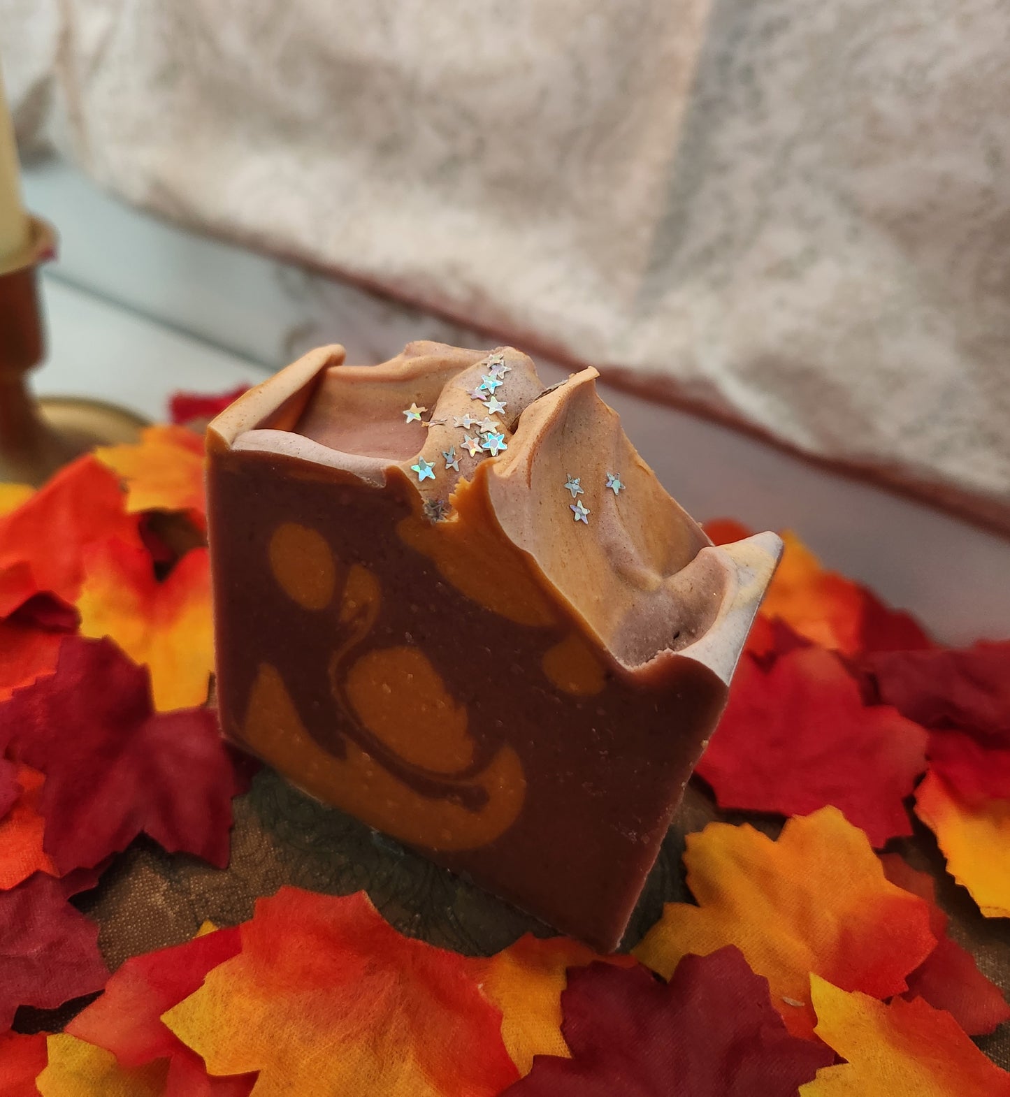 A handmade rectangular bar of soap with a swirled red and orange design, decorated with small silver glitter stars, displayed on a bed of colorful autumn maple leaves.